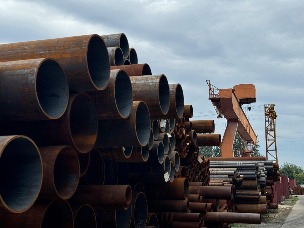 Главная страница Rusty steel pipes stacked outdoors with cranes and cloudy sky.